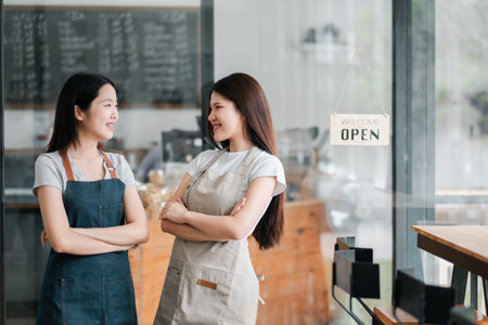 Two friendly baristas in a modern cafe, wearing aprons, standing with arms crossed, smiling at each other near an open sign.の写真素材