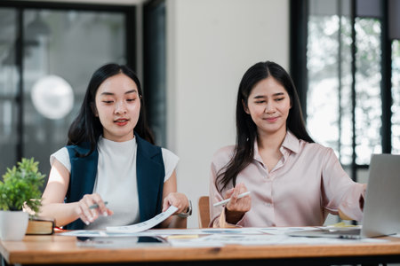 Two women working together in a contemporary office, reviewing documents and using a laptop.の写真素材