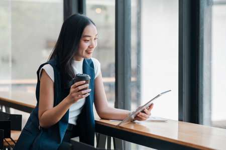 Smiling young woman holding a coffee cup and using a tablet in a bright, modern office setting.の写真素材