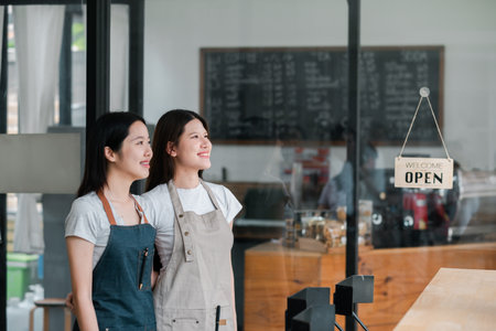 Two women in aprons stand inside a cafe, looking outside with smiles, next to an open sign on the glass door.の写真素材