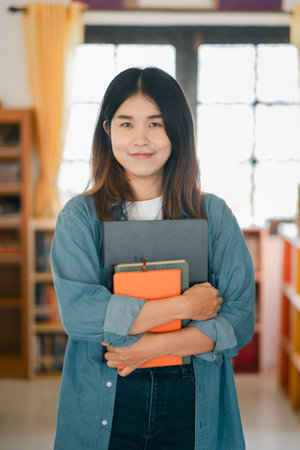 Smiling young woman holding books in a cozy library with warm lighting and wooden shelves.の写真素材