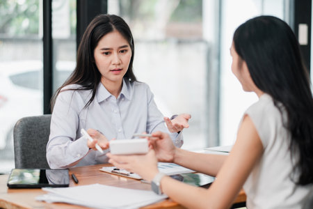 Two women engaged in a business discussion at a modern office, using digital devices and documents.の写真素材