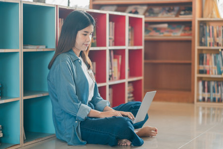 A young woman sits on the floor using a laptop in a modern library with colorful bookshelves, focused on study or work.の写真素材