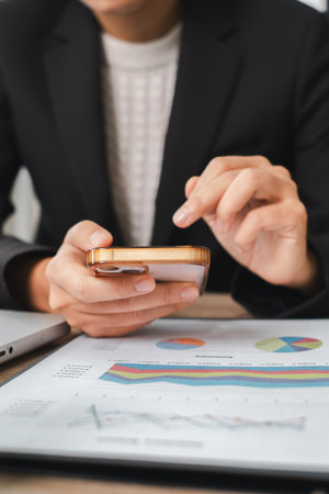 Close-up of a business professional using a smartphone to analyze financial data with charts and graphs in a modern office.の写真素材