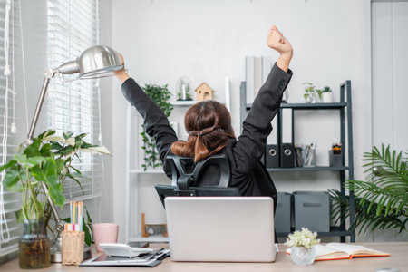 A joyful businesswoman in a suit celebrates success at her desk in a modern office, surrounded by greenery and office supplies.の写真素材
