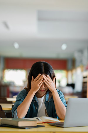 A student sitting at a desk in a library, holding their head in frustration while surrounded by books and a laptop.の写真素材