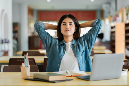 Young woman in denim shirt sitting at a desk with a laptop, looking thoughtful in a library...