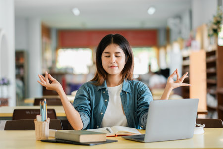 A young woman meditates at a desk in a library, surrounded by books and a laptop, embodying mindfulness and relaxation in a modern educational setting.の写真素材