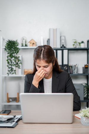 A businesswoman in a black blazer appears stressed while sitting at a desk with a laptop in a modern office environment.の写真素材