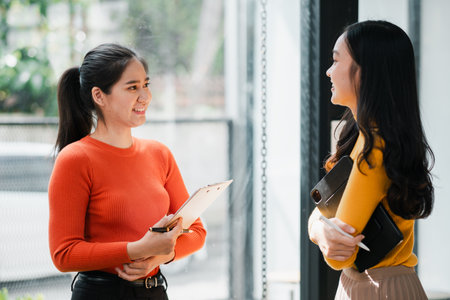 Two women chatting in a bright office, wearing casual clothes, holding notebooks, and smiling warmly.の写真素材