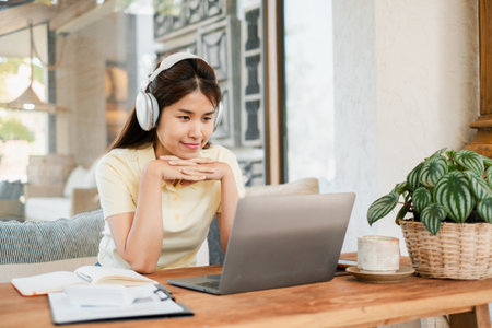A young woman wearing headphones, focused on her laptop in a cozy home office setting with natural light and greenery.の写真素材