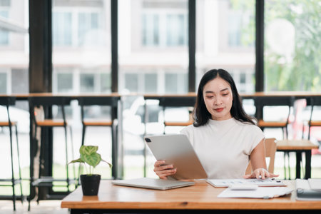 A young woman using a tablet in a bright, modern setting with large windows, focusing on her work.の写真素材