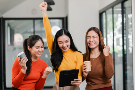 Three young women cheerfully celebrate success in a modern office, holding a tablet and coffee cups.