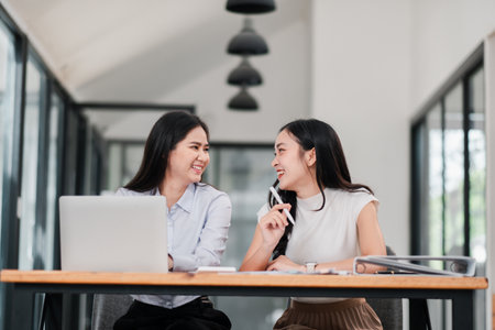 Two women smiling and talking at a desk in a modern office setting, showcasing teamwork and collaboration.の写真素材