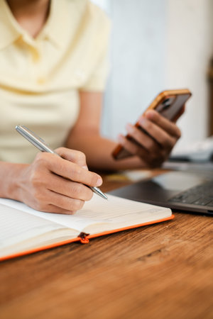 Close-up of a person writing in a notebook while holding a smartphone, with a laptop on a wooden...