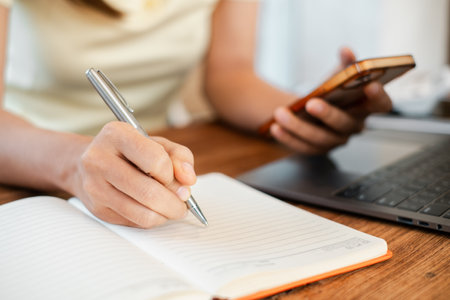Close-up of a person writing in a notebook while holding a smartphone, with a laptop nearby on a wooden desk.の写真素材