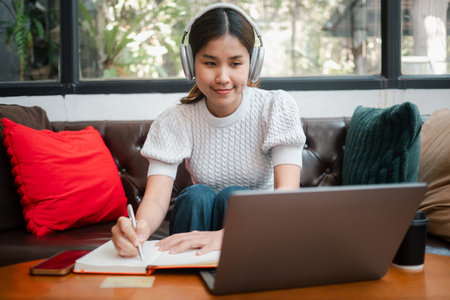A young woman wearing headphones studies on her laptop while taking notes in a cozy living room...