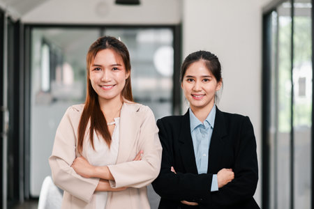 Two professional women in business attire confidently pose with arms crossed in a bright, modern office setting.の写真素材