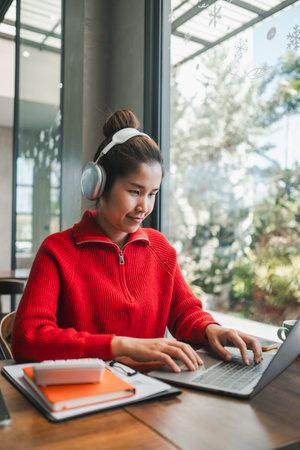 Smiling woman wearing headphones, working on a laptop in a bright, modern cafe with large windows...