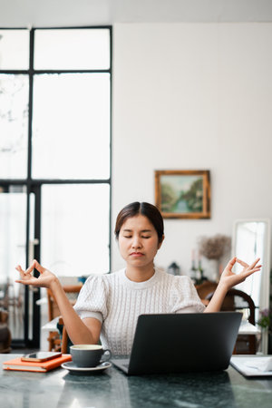 Woman meditating at home office desk with laptop and coffee, embracing mindfulness in a modern interior setting.の写真素材