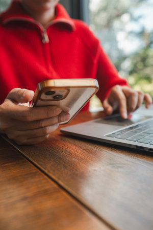 Close-up of a woman multitasking with a smartphone and laptop, wearing a red sweater, indoors.の写真素材