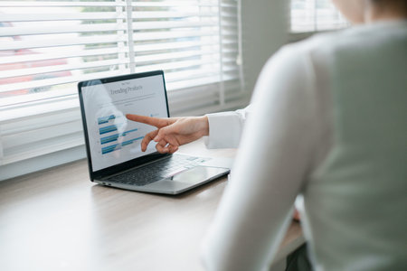 A business professional points at a laptop screen displaying data charts, analyzing trends in a...