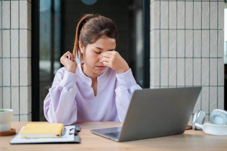 Young woman feeling stressed at work, sitting at a desk with a laptop and documents in a modern office environment.の写真素材