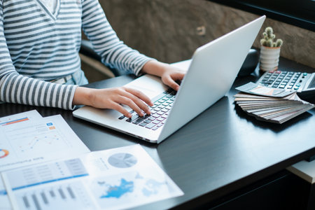 A person typing on a laptop with financial documents and a calculator on a desk in a modern office environment.の写真素材