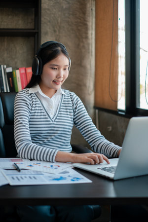 A young woman wearing headphones works on a laptop in a modern office, surrounded by documents and books.の写真素材