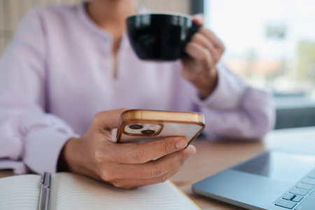 Close-up of a woman holding a smartphone and coffee cup, working at a cafe with a laptop and...