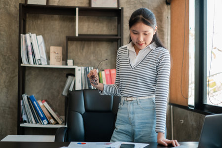 Young woman in a modern office reviewing business documents, standing by a desk with a bookshelf in the background.の写真素材