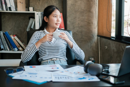 Young woman in striped shirt taking a coffee break in a modern office setting with documents and a laptop.の写真素材