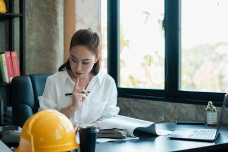 A focused female architect examines blueprints in a contemporary office setting, highlighting...