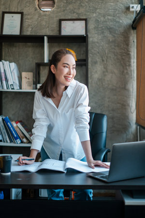 Young woman in a white shirt working at a desk with a laptop and documents, smiling confidently in a modern office.の写真素材
