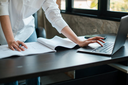 A professional in a white shirt working on a laptop and reviewing documents in a contemporary office...