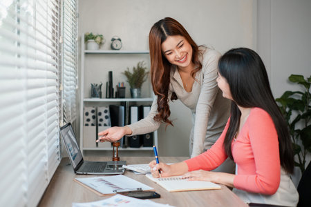 Two women working together in a bright office, discussing ideas and using a laptop, showcasing teamwork and productivity.の写真素材