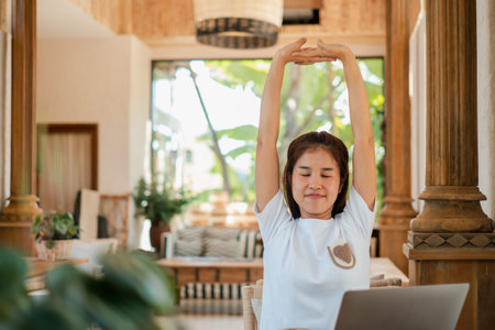 A young woman stretches while working on her laptop in a cozy, naturally lit room with wooden decor...