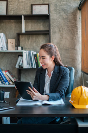Professional woman in office setting using a tablet, with a safety helmet on the desk, symbolizing industry and technology.の写真素材