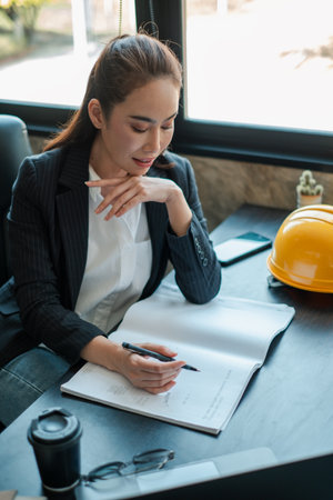 Professional woman in office setting reviewing documents, with a hard hat and coffee cup on the desk, symbolizing work-life balance.の写真素材