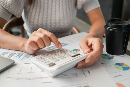 Close-up of hands using a calculator on financial charts, with a coffee cup nearby, in a modern office environment.の写真素材