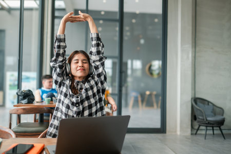 Woman in plaid shirt stretching with headphones on, sitting at a laptop in a modern cafe setting.の写真素材