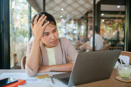 Young woman looking stressed while working on a laptop in a modern cafe setting, surrounded by...