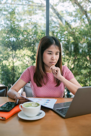 Young woman concentrating on laptop work in a cafe, surrounded by documents and coffee, with a natural backdrop.の写真素材