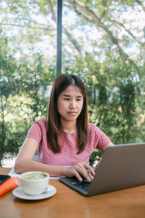 Woman in a pink shirt working on a laptop in a cafe with a coffee cup, surrounded by greenery.
