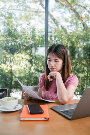 A woman in a pink shirt reviews documents at a cafe table, with a laptop, coffee, and notebook...
