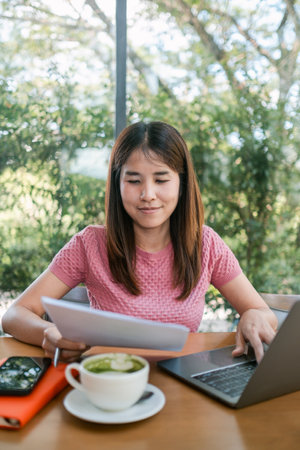 A focused young woman in a cafe, working on a laptop with documents, enjoying a coffee, surrounded by greenery.の写真素材