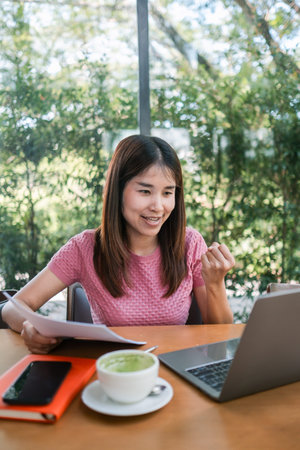 Smiling woman working on a laptop in a cafe, surrounded by greenery, with a cup of coffee and...