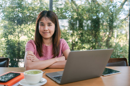 A young woman sits at a table with a laptop and a cup of coffee, working remotely in a cafe surrounded by lush greenery.の写真素材