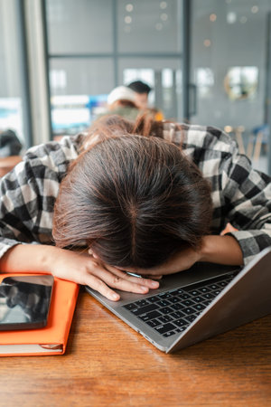 Tired woman in plaid shirt resting on laptop at a cafe table, surrounded by digital devices,...