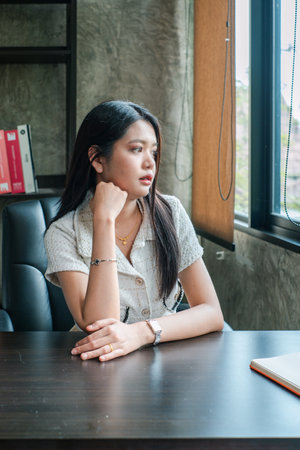 Young woman in a modern office, sitting at a desk, gazing thoughtfully out the window.の写真素材